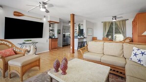 A spacious living room with a sofa, a coffee table, and a TV mounted on the wall. There is a kitchen area visible in the background. - Ocean & Golf Course View Condo at Keauhou Akahi | 1 Bedroom + Murphy Bed (Kailua Kona)