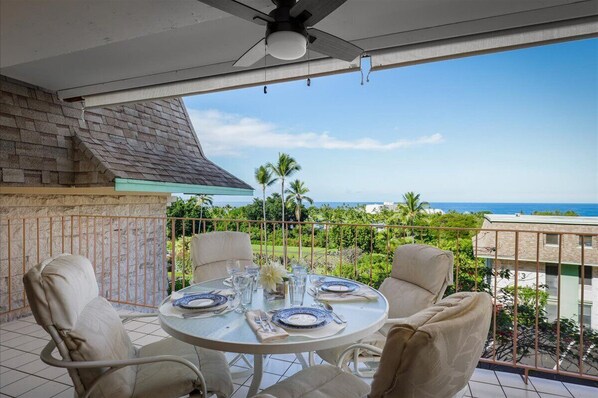 Un balcón con una mesa dispuesta para comer, con vistas a la costa, palmeras y un edificio al fondo.