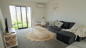 A living room with a grey sofa, a small round table with chairs, a TV on a stand, and a large sliding glass door leading to a garden.