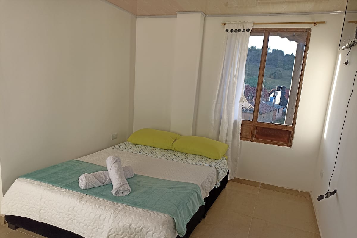 Uma cama de solteiro com colcha branca e azul-turquesa, um travesseiro verde e uma toalha enrolada sobre a cama. Há uma janela com moldura de madeira e cortinas brancas, e uma vista de edifícios e árvores lá fora.