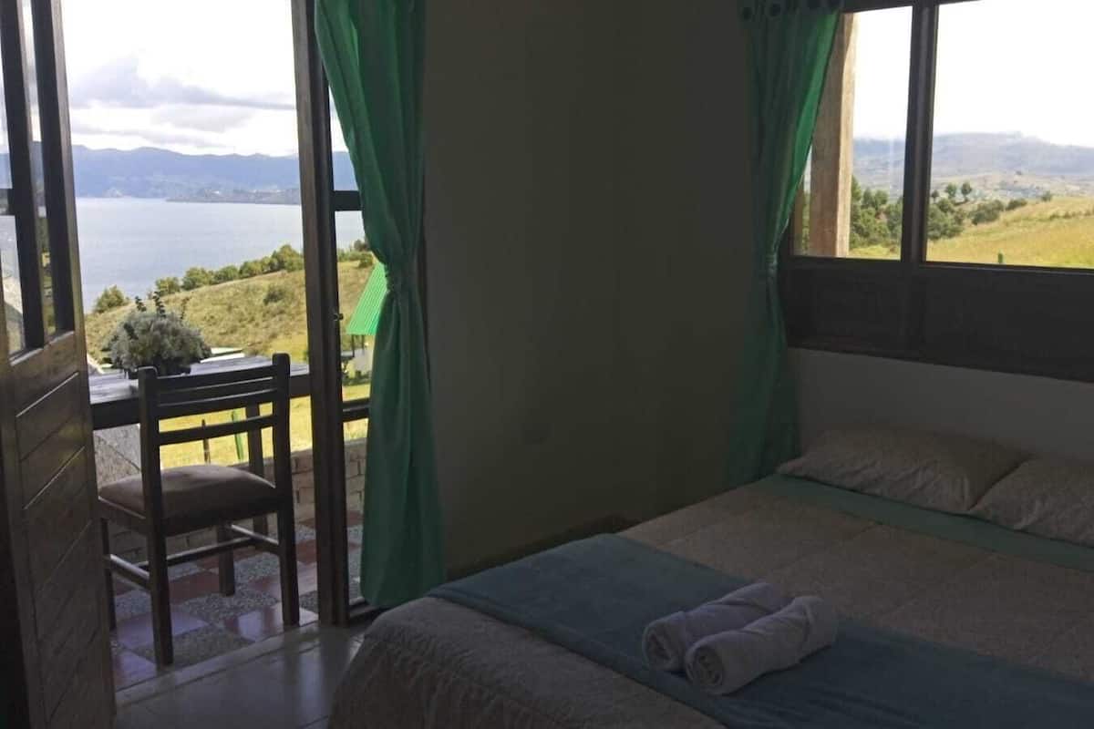 Quarto com cama, uma cadeira e vista para um lago e colinas.