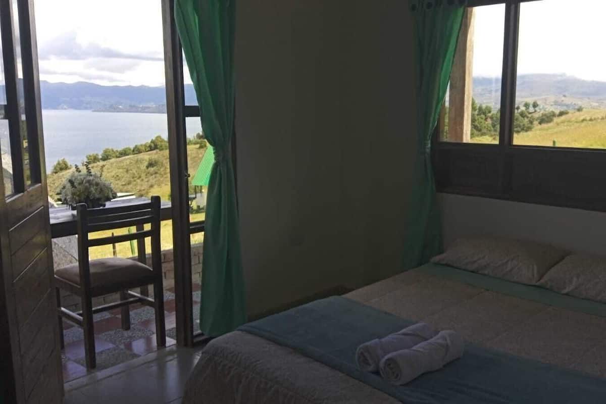 Quarto com cama, uma cadeira e vista para um lago e colinas.