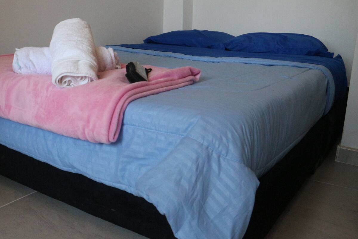 Cama com manta azul e rosa, toalha enrolada e um pequeno objeto preto sobre a cama.