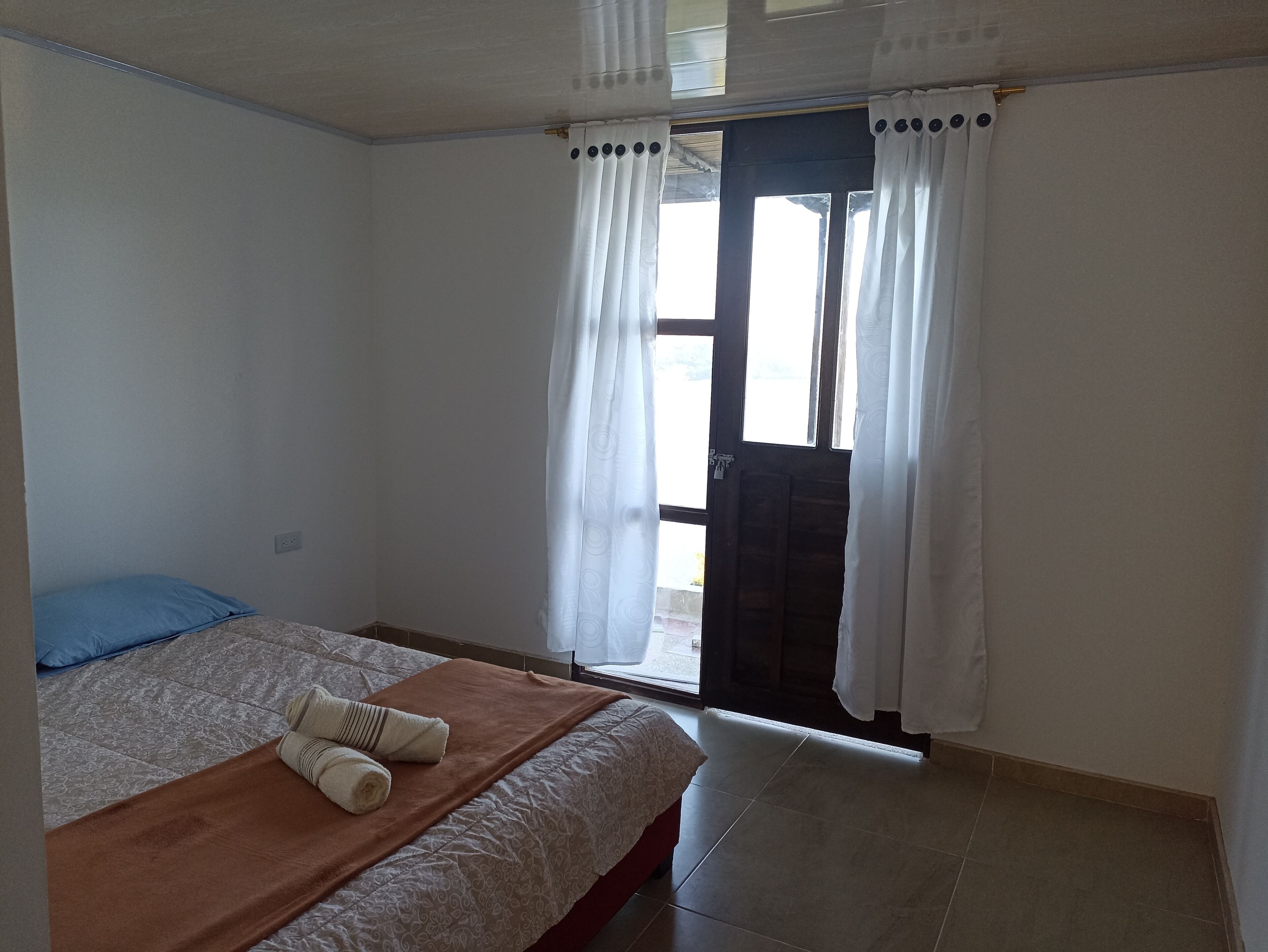 A bedroom with a bed, a blue pillow, a brown blanket, and a rolled towel. There is a door with a curtain and a view of the outside.