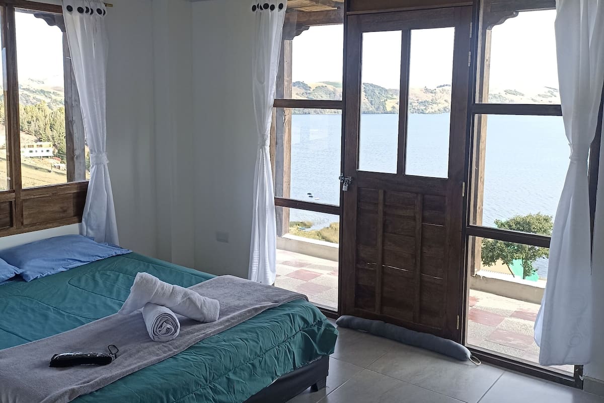 Quarto com cama, janela com cortinas e vista para o mar.