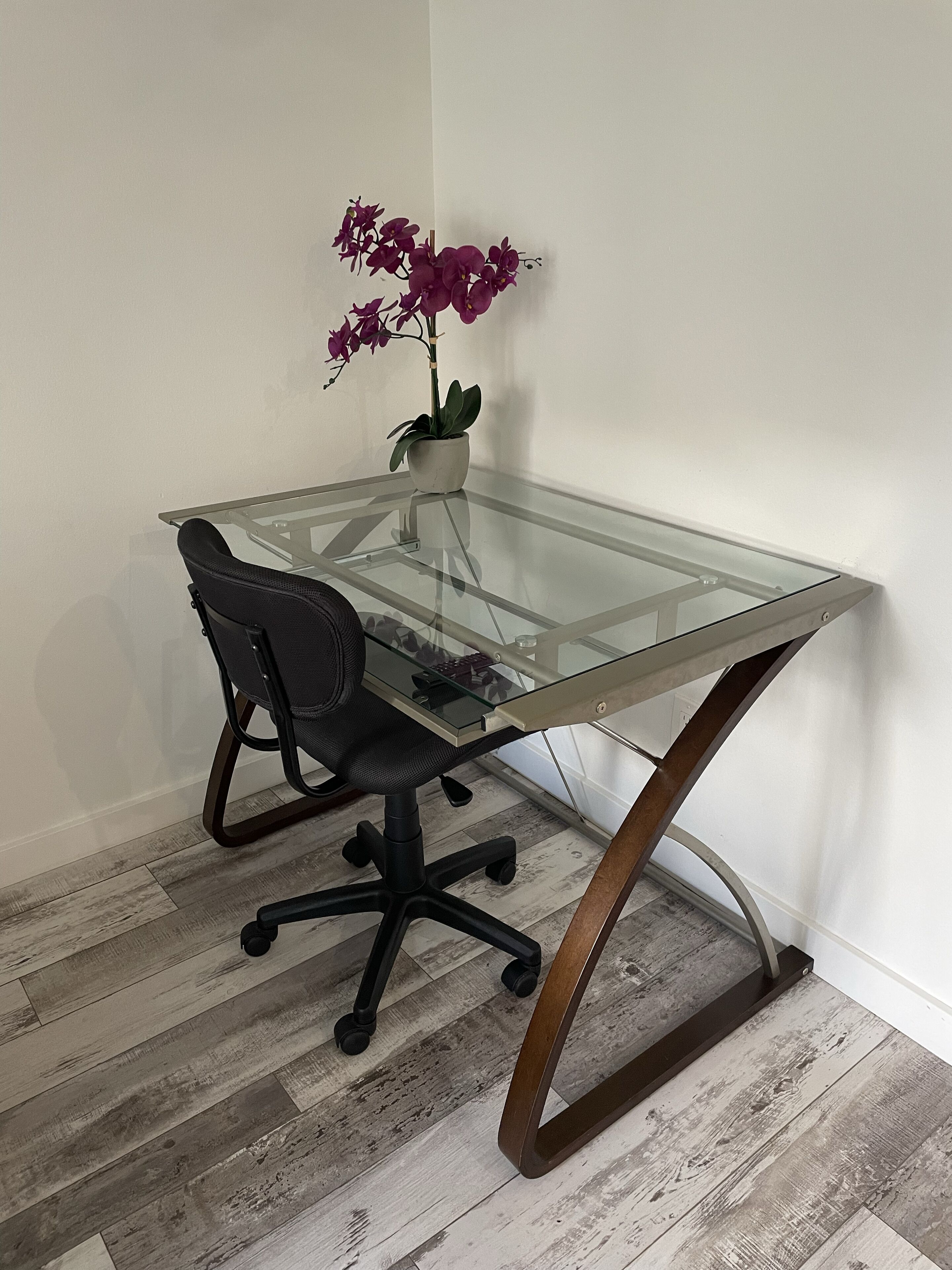 Un bureau à plateau en verre, une chaise de bureau noire, une orchidée en pot et un plancher en bois.
