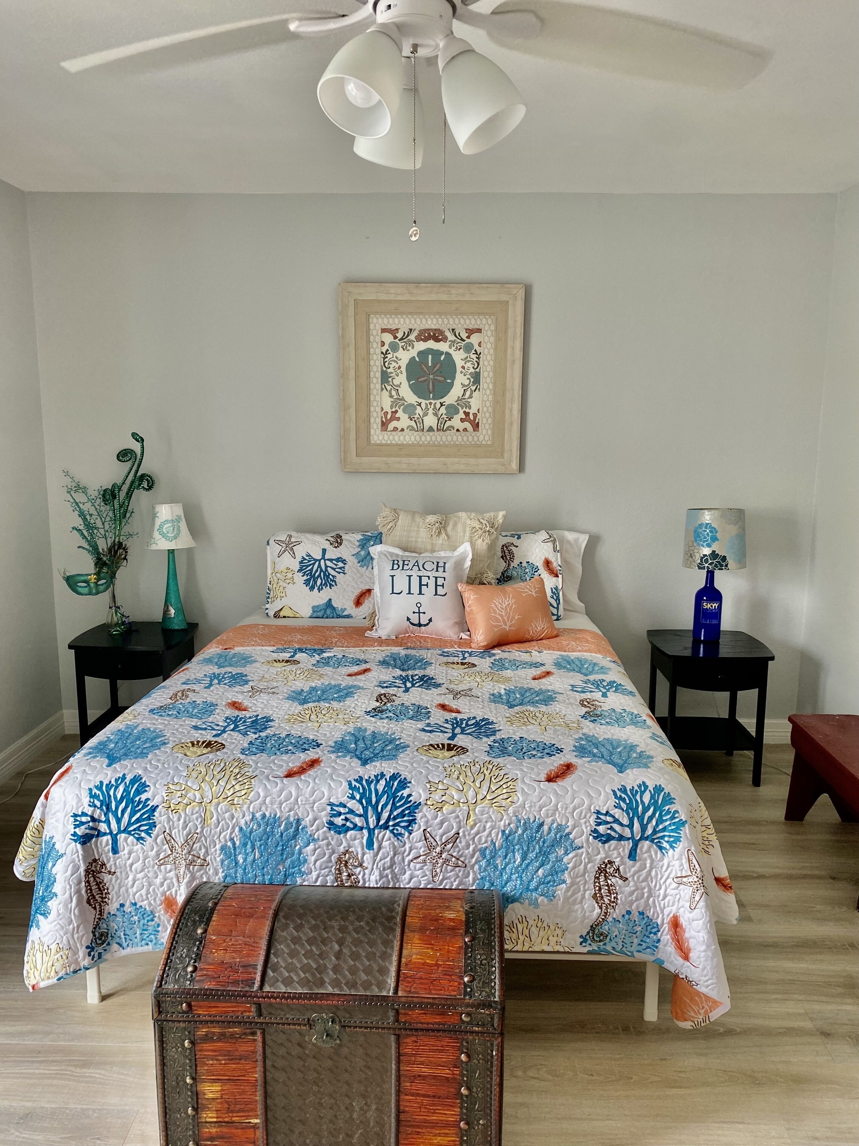 Un dormitorio con una cama, un ventilador de techo, una obra de arte enmarcada, una mesita de noche, una lámpara, una planta decorativa y un aparador.