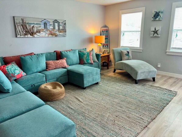 A living room with a teal sectional sofa, a round ottoman, a woven rug, a lamp, and wall art including a welcome sign and marine-themed pieces.