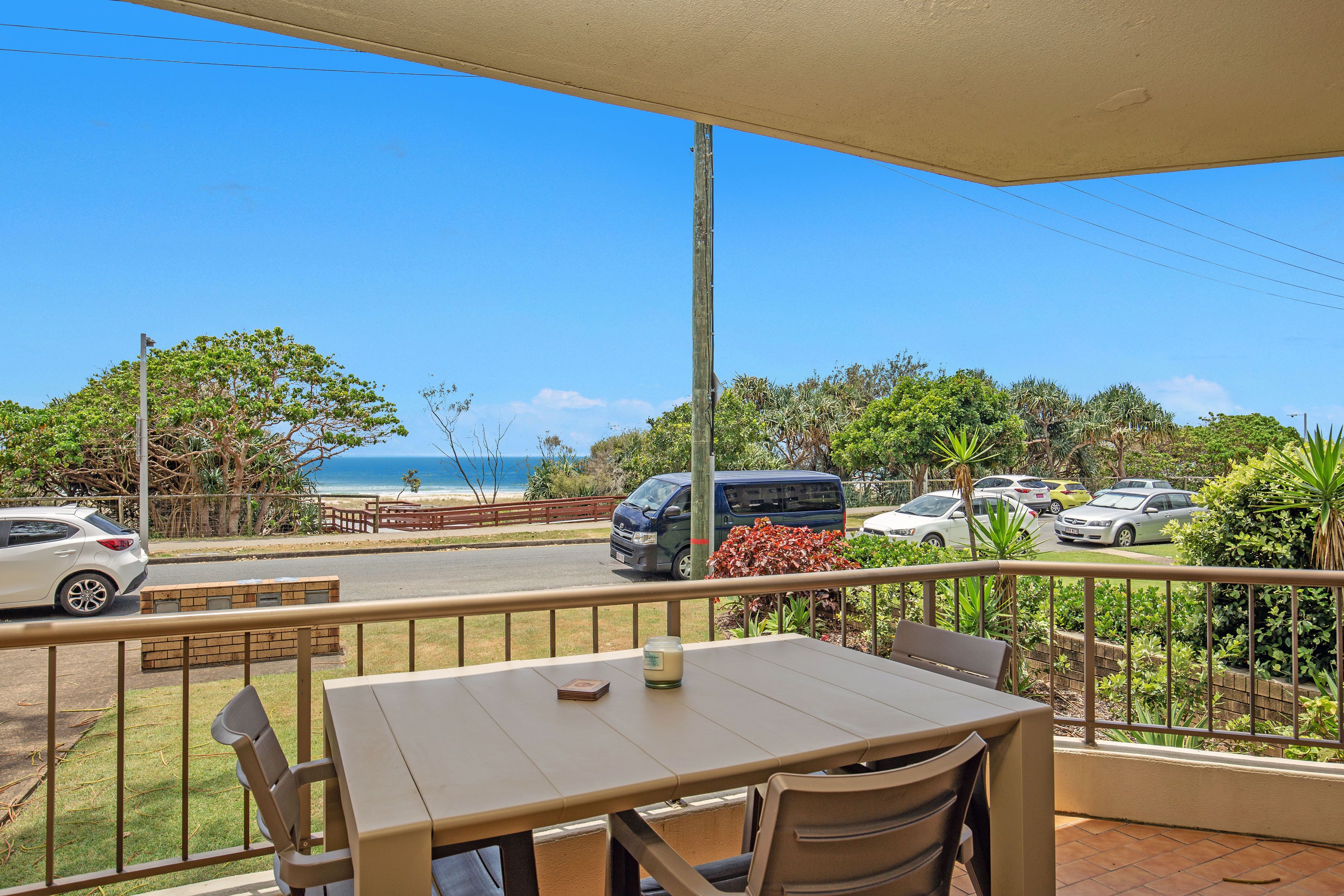 A balcony with a table and chairs, overlooking a parking area and a beach.