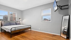 A bedroom with a bed, two windows, a wall-mounted TV, and a mirror.