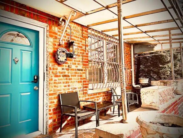 Guests enter through a distinctive blue door, accessing a room with a brick wall and metal gate, a seating area with a bench and table, and a large window with a metal frame.