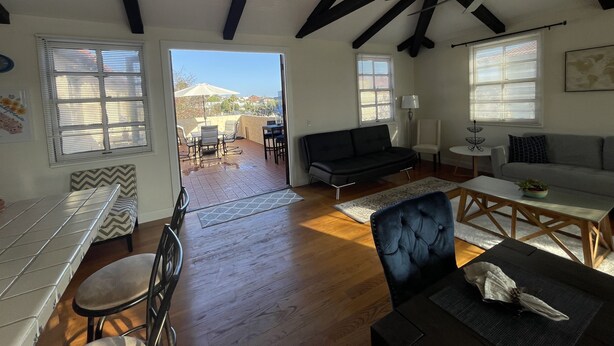 A spacious living area with a wooden floor, a black sofa, a dining table, and a view of the outdoors through large windows. - San Diego Old Town Family-sized Apartment with Patio (San Diego)