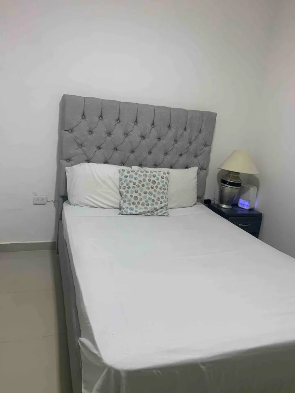 A bed with a tufted headboard, a bedside lamp, and a power outlet on the wall.