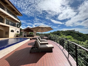 Relax by the pool on a spacious deck surrounded by lush greenery and a hilly landscape.