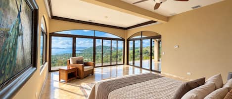 A spacious bedroom with a large bed, a ceiling fan, and a view of the outdoors through large windows.