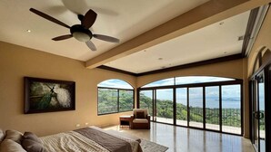 A spacious bedroom with a large bed, a ceiling fan, a painting, and a balcony with a view of the landscape. - Stunning Ocean View Chateau - Experience it All (Playa Hermosa, Sardinal)