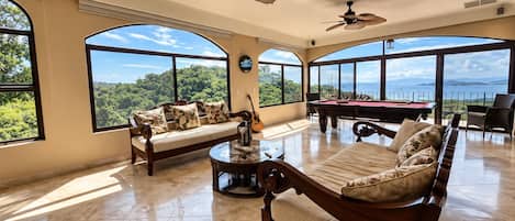 A spacious living room with a pool table, two sofas, and large windows offering a view of the outdoors.