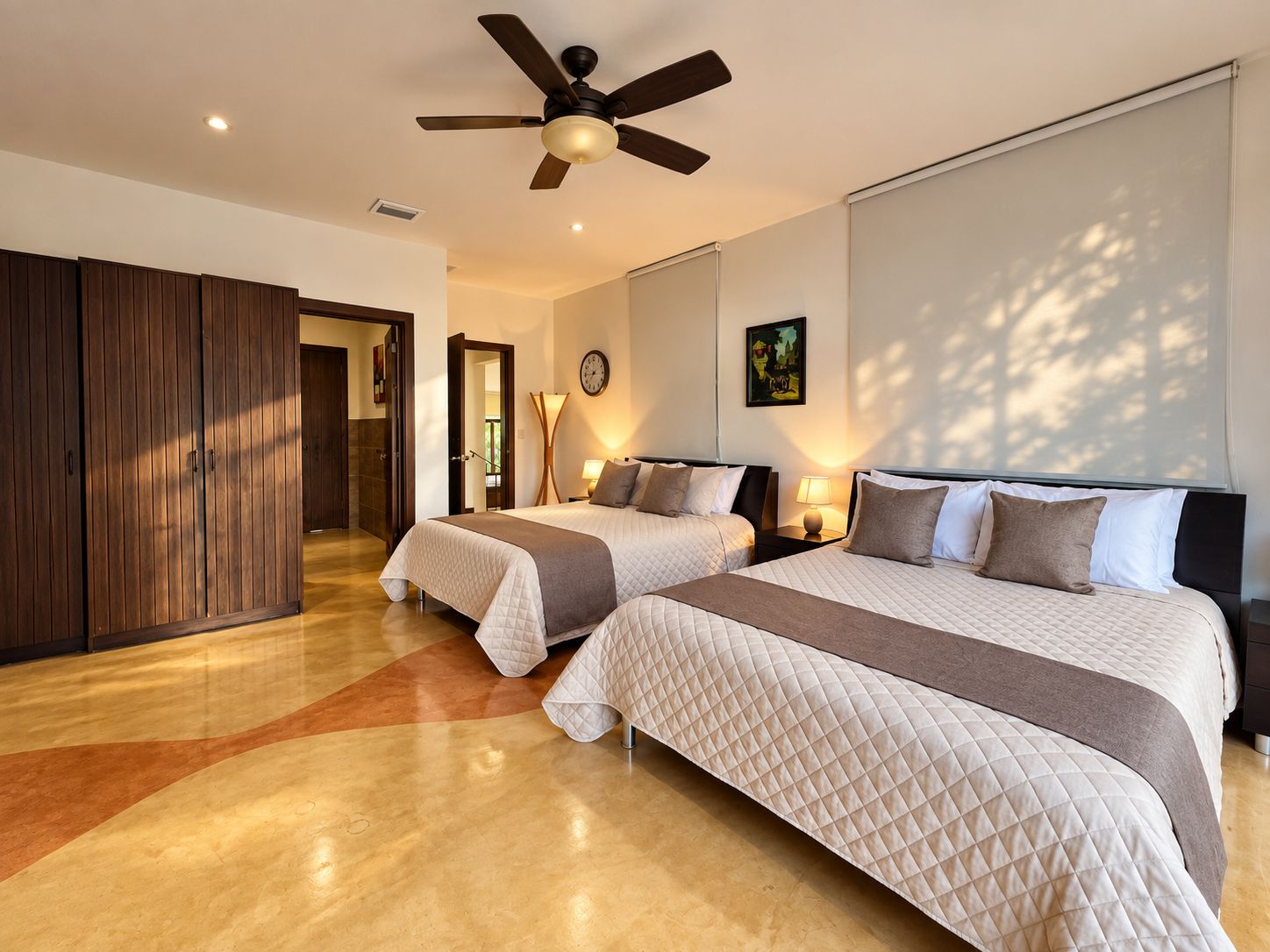A hotel room with two beds, a ceiling fan, a painting on the wall, and a wooden wardrobe.