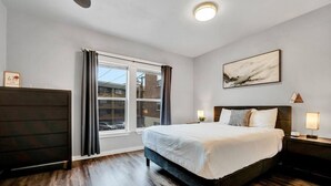A hotel room with a large bed, a dresser, a window with curtains, a painting on the wall, and a nightstand with a lamp. - New! Prime DT Modern - Extended stay Equipped! (Tacoma)