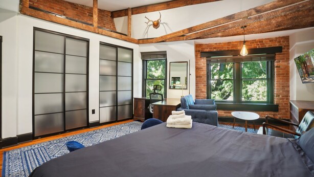 A bedroom with a large bed, a desk, a chair, a small table, and a window with a view of trees. - 8BR/7BA Mansion · Sleeps 28 · Shaw Metro (Washington)