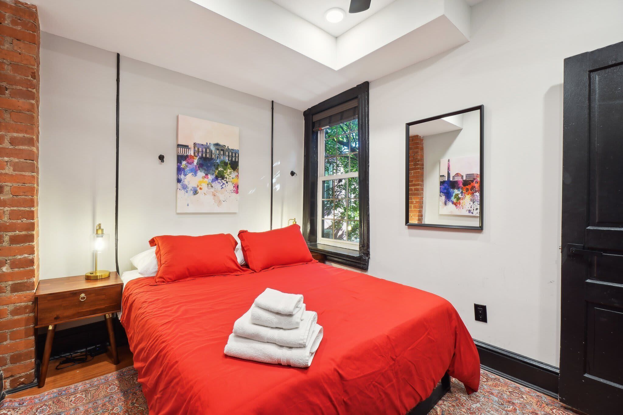 Quarto de hotel com colcha vermelha, mesa de cabeceira com abajur, janela com vista para árvores e porta.