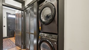 A laundry room with two stacked front-loading washing machines, a door leading to another room, and a wooden floor. - 8BR/7BA Mansion · Sleeps 28 · Shaw Metro (Washington)