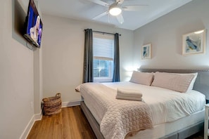 A bedroom with a bed, a ceiling fan, a TV, a window with curtains, and a wall-mounted artwork.