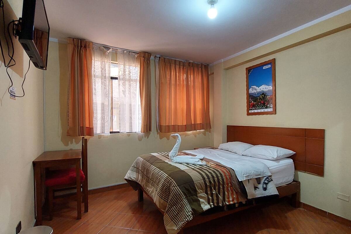 A bedroom with a wooden bed, a desk, a chair, and a television mounted on the wall.