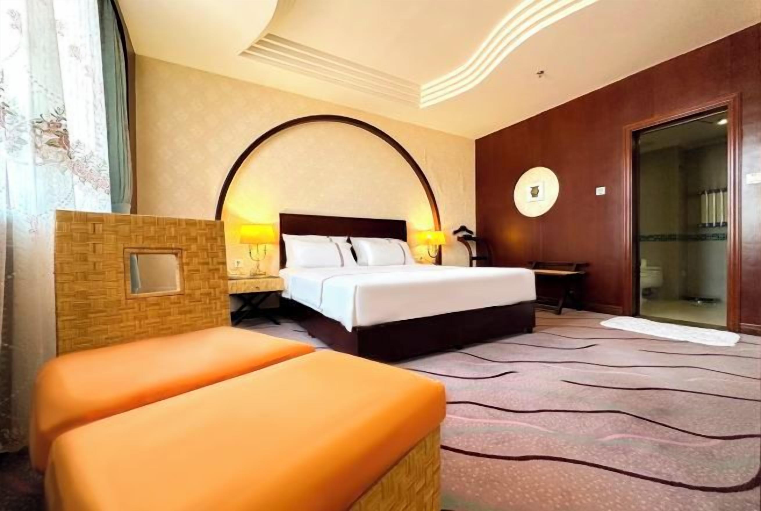 A hotel room with a bed, a small table, a chair, a mirror, and a large window with curtains.