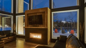 Ein modernes Wohnzimmer mit Kamin, Fernseher und großen Fenstern mit Blick auf eine verschneite Landschaft.