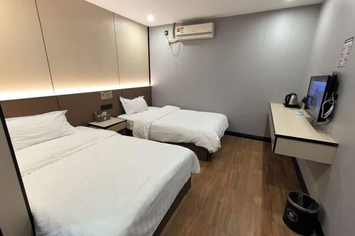 A hotel room with two beds, a bedside table, a desk with a computer, and an air conditioning unit.