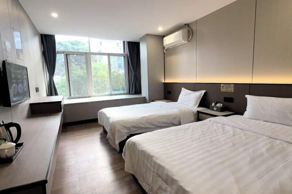 A hotel room with two beds, a TV, a window with curtains, and a wall-mounted air conditioner.