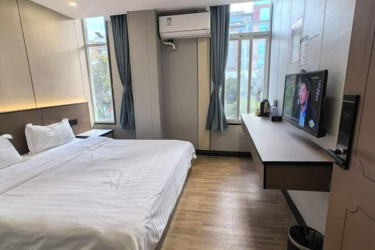 A hotel room with a bed, a desk with a television, an air conditioning unit, and a window with curtains.