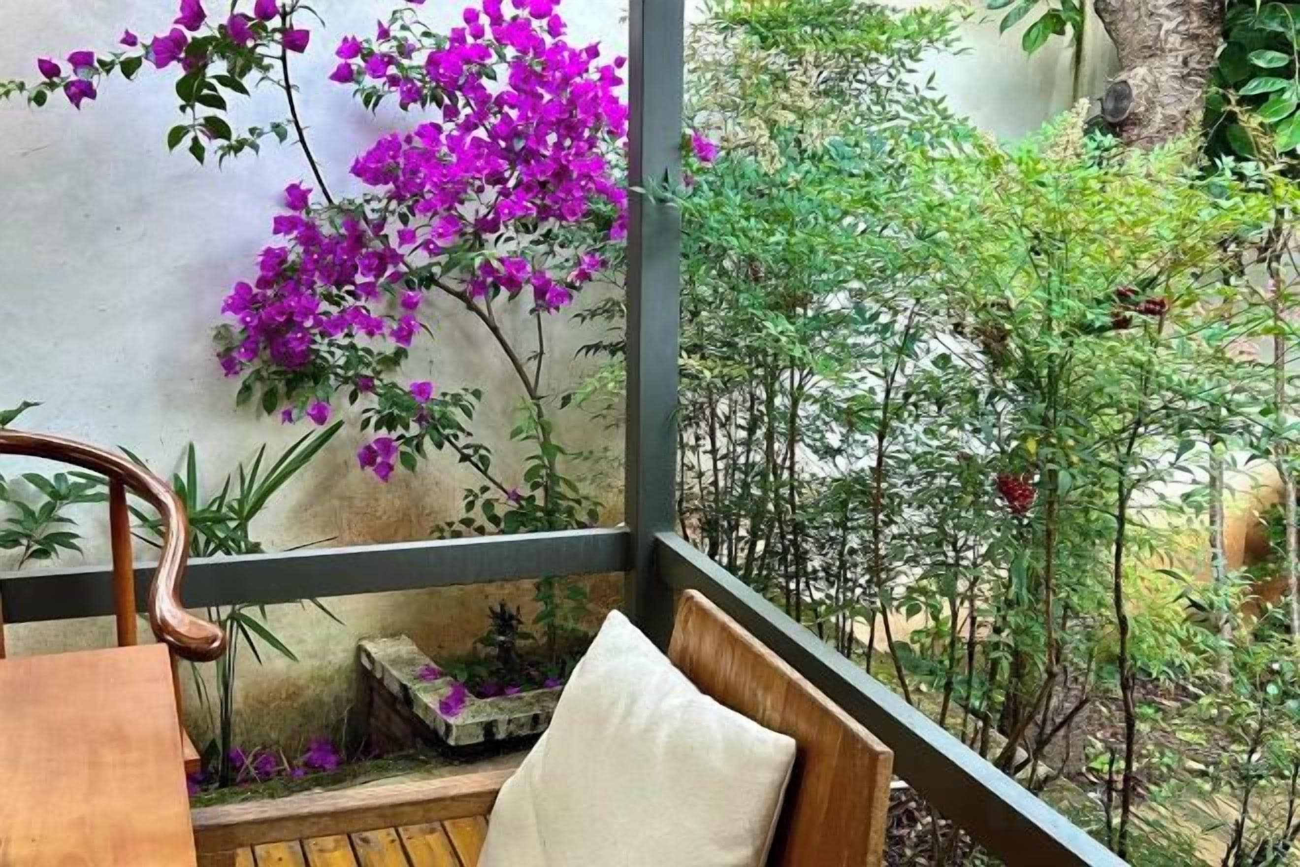 A balcony with a wooden table, a cushioned bench, and a view of a garden with purple flowers and greenery.