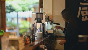 Um barista prepara uma bebida em uma máquina de café em um café com vista para o verde lá fora.