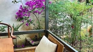 Uma varanda com uma cadeira de madeira, uma mesa pequena e vista para um jardim com flores roxas e folhagem verde.