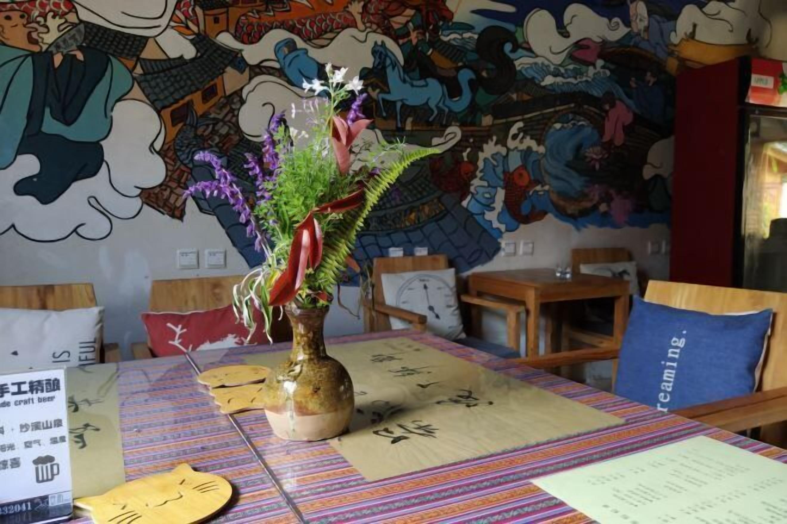 A dining area with a table set for two, featuring a vase with flowers and a menu. The wall behind has a colorful mural.