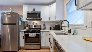 Una cocina moderna con electrodomésticos de acero inoxidable, armarios blancos, horno empotrado y una ventana con persianas.