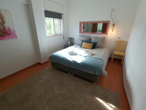 Une chambre à coucher comprenant un lit, une table de chevet, une chaise, une fenêtre avec des stores et un miroir fixé au mur.