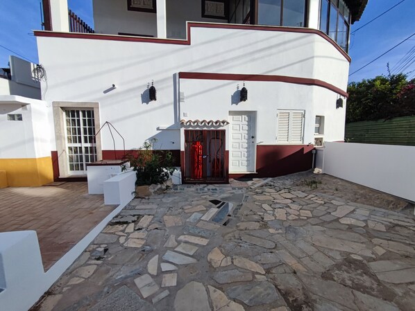 A white building with a stone-paved courtyard, a red door, and a small garden area.