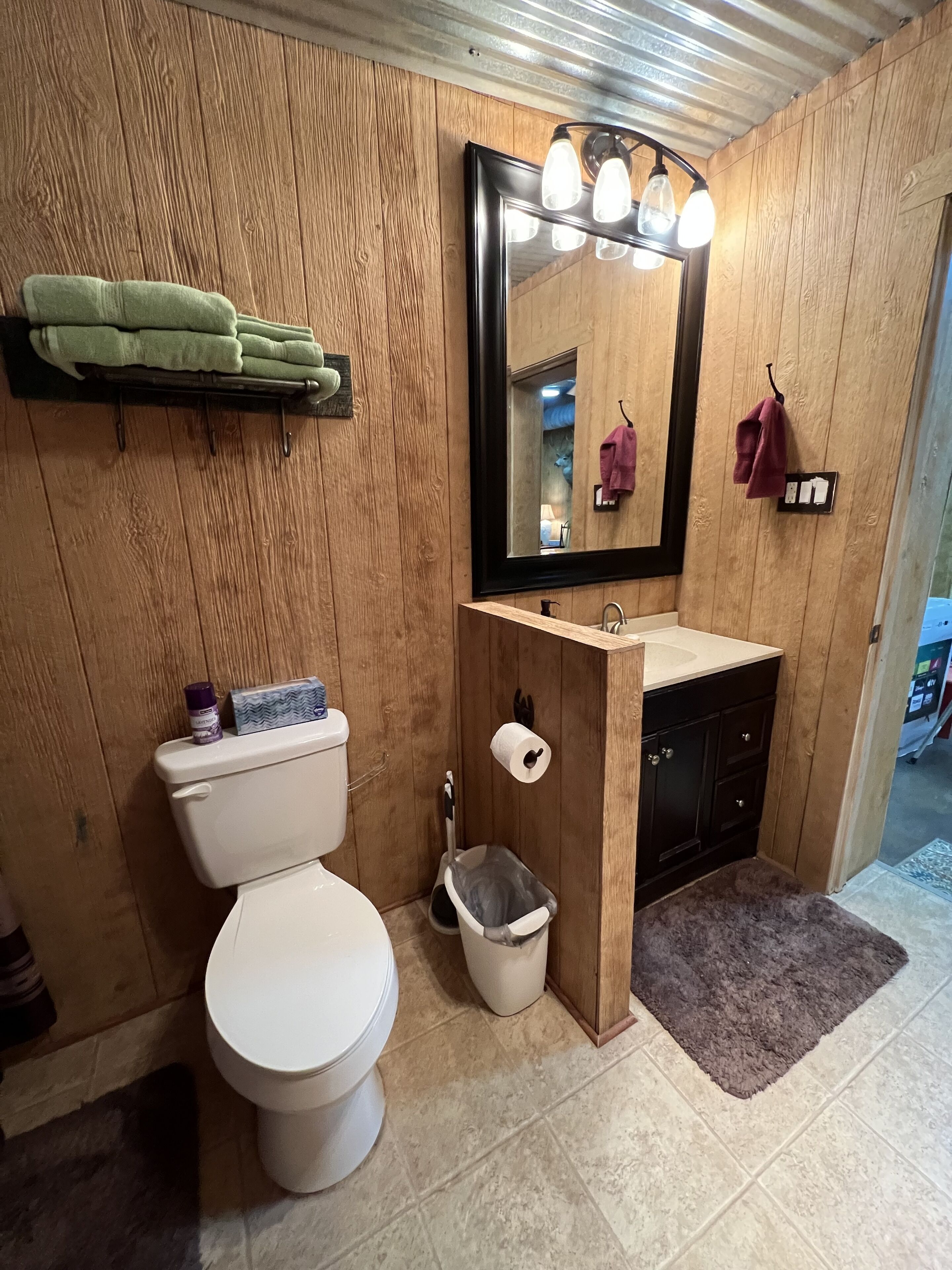 Un baño pequeño con pared de madera, inodoro blanco, lavamanos con espejo y un pequeño cubo de basura.