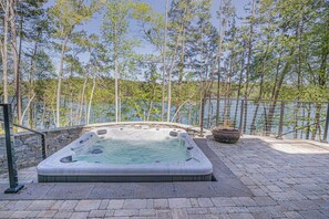 A hot tub surrounded by a stone wall, a metal railing, and a large fire pit, set against a backdrop of trees and a body of water.