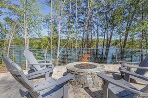 An outdoor seating area with chairs and a stone fire pit, overlooking a lake with trees in the background.