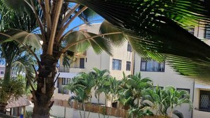 A poolside view with a palm tree, a wooden deck, and a building with multiple windows. - Sugareef 3-Bed Apartment in Flic en Flac (Flic en Flac)
