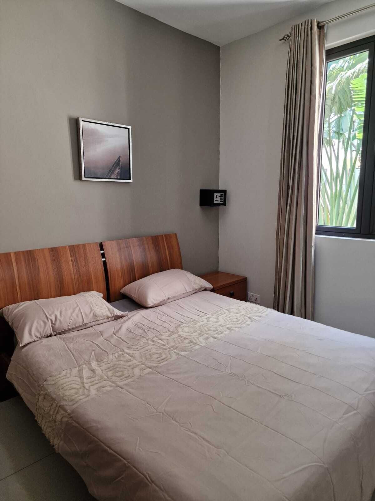 Un dormitorio con cabecera de madera, cama con colcha estampada, mesita de noche, termostato en la pared y ventana con cortinas.