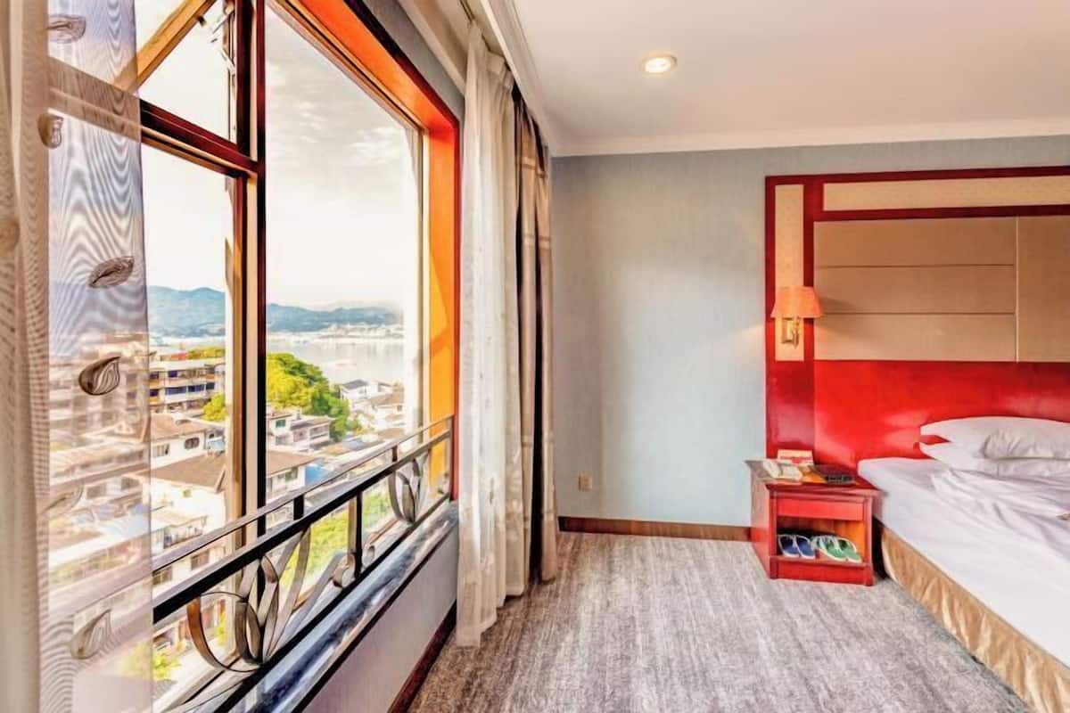 A hotel room with a large window, a bed with a red headboard, a nightstand, and a view of the cityscape.