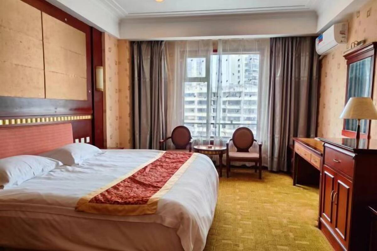 A hotel room with a bed, a desk, a chair, a window with curtains, and a view of buildings.