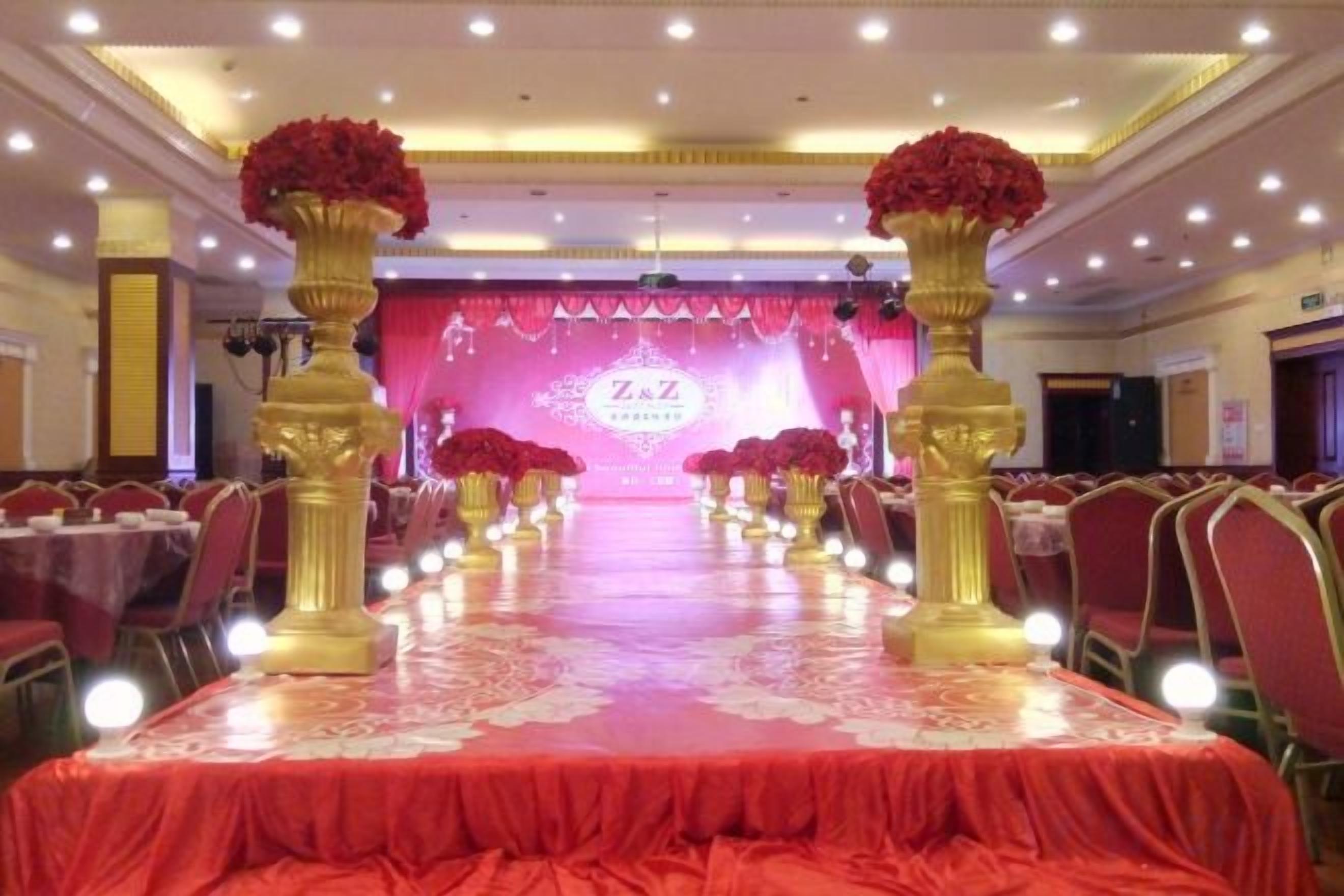 Un salón de banquetes con mesas dispuestas en filas, un escenario con fondo rosa y pilares dorados adornados con flores rojas.