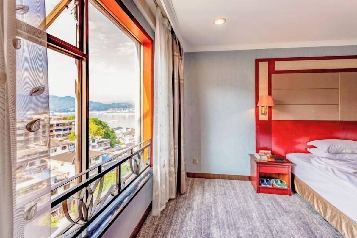 A hotel room with a large window, a bed with a red headboard, a nightstand, and a view of the city.