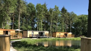 Een rij houten hutten met een vijver op de voorgrond, omringd door bomen.